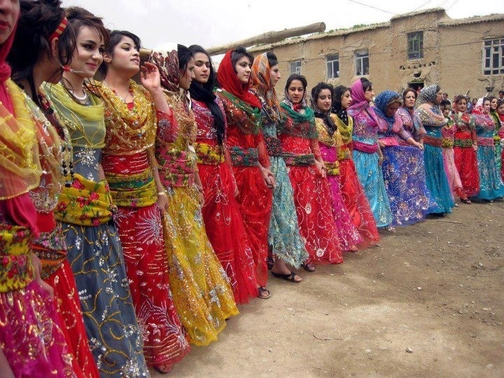 Kurdish women's traditional dress — layered silk and velvet robes in deep jewel tones, with embroidered cuffs, coin headdresses, and a woven sash at the waist